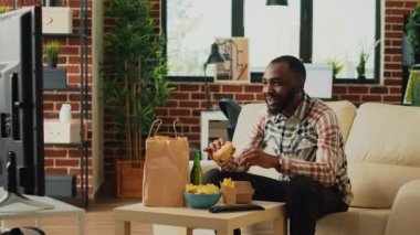 Cheerful man taking bite of hamburger in living room, eating fast food from takeaway delivery order. Happy adult watching television at home and serving burger with fries and alcohol.