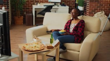 Young woman holding bowl of chips watching tv, drinking beer and eating delivery meal from multiple fast food orders. Female adult having hamburgers, fries, pizza and noodles on table.