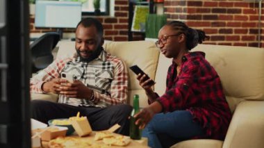 African american couple browsing apps on smartphones, using online internet on mobile phones and watching television together. Young modern partners using phones, takeaway food.