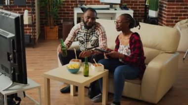 Happy life partners eating asian meal from delivery, serving noodles with chopsticks and watching comedy movie on tv. Modern couple in relationship enjoying show and food with alcohol.