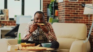 African american man eating slice of pizza at home, binge watching action tv series in living room. Modern happy guy enjoying fast food delivery and having fun with movie. Tripod shot.