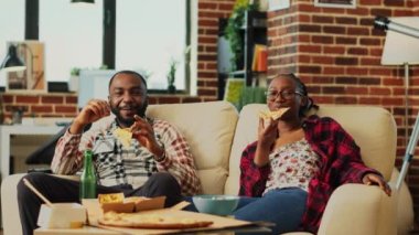 Young couple eating slices of pizza and watching show on television, laughing and being together. Man an woman in relationship enjoying takeaway meal to watch favorite film on tv. Tripod shot.