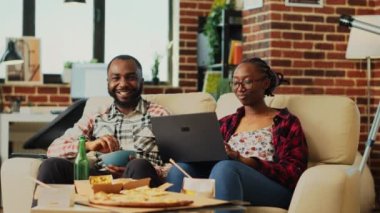 Young people in relationship watching tv show at home, eating snacks and using laptop to browse online app. Man and woman having fun with movie or film, relaxing at television. Tripod shot.