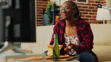 Smiling woman eating pizza slice in living room, enjoying comedy movie on television. Young person feeling happy drinking bottle of beer and having dinner from takeaway delivery. Handheld shot.