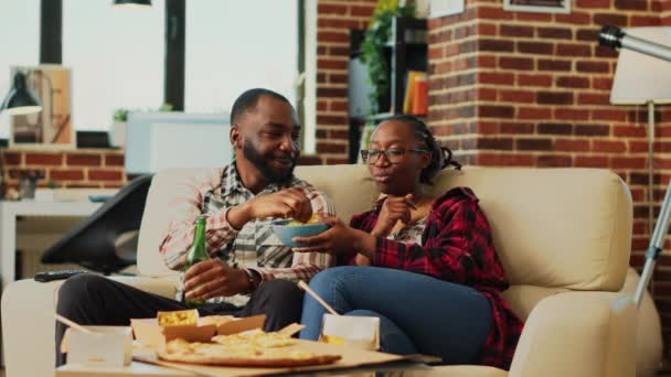 非洲裔美国人夫妇看电视和吃薯片 在电视上轻松地喝着啤酒 男朋友和女朋友喜欢看动作片 吃喝玩乐 三脚架射击 — 图库视频影像