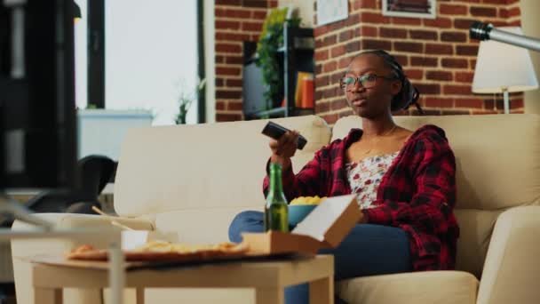 非洲裔美国女孩打开电视看电影 吃薯片和在沙发上喝酒 微笑的女人对电视前的快餐餐感到开心 手持射击 — 图库视频影像