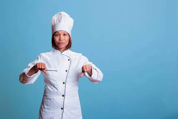 Elderly cook woman wearing kitechen uniform doing disapproval sign after looking at culinary dinner meal. Cheerful chef showing thumbs down gesture, preparing gastronomy healthy dish