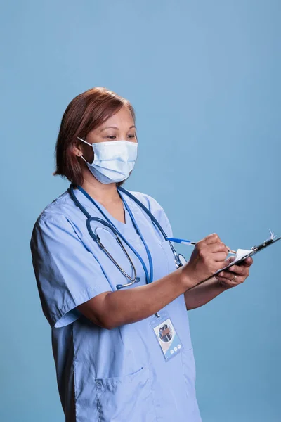 Elderly asian nurse writing medical expertise on clipboard analyzing disease diagnosis during checkup visit appointment. Assistant wearing protective face mask to prevent coronavirus