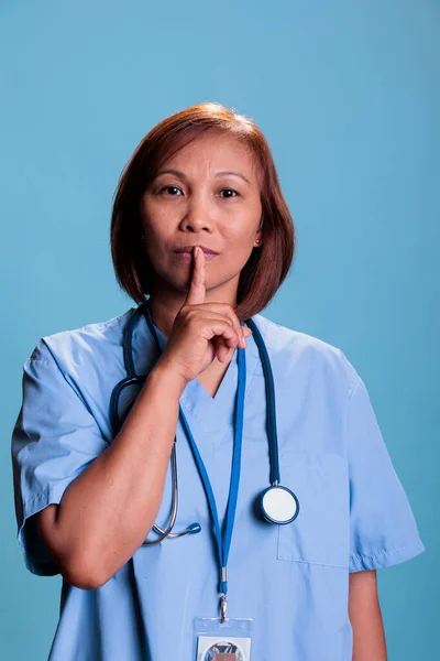 Old physician nurse showing shhh taboo sign with finger to lips while working at health care expertise during checkup visit appointment. Specialist assistant wearing blue uniform and stethoscope.