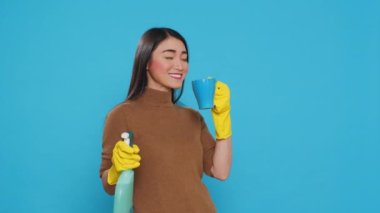 Happy housekepper enjoying cup of coffee while cleaning house with chemical spray bottle, standing in studio over blue background. Maid using protective equipment and sanitary practices