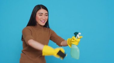 Happy asian maid wearing protective gloves while cleaning house using sponge and detergent spray, standing in studio over blue background. Housekeeper is an expert in cleaning equipment