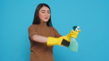 Cheerful asian housewife wearing rubber gloves while cleaning house using sponge and detergent spray, standing in studio over blue background. Smiling housekeeper is an expert in cleaning