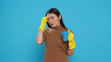 Pensive thoughtful maid holding cup of coffee standing over blue background in studio, thinking at solution to solve cleaning problem. Housekepper providing top-notch cleaning services to her clients.