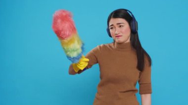 Exhausted sleepy housekeeper wearing headphones while doing cleaning in house using feather duster, yawning in front of camera. Overwhelmed maid role was vital to the cleanliness of many homes