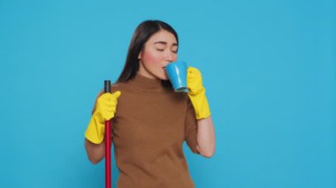 Cheerful housekepper enjoying cup of coffee while cleaning house with broom, standing in studio over blue background. Maid using protective equipment and sanitary practices to keep customers safe.