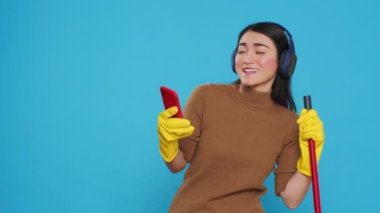 Happy cleaning lady wearing headphone while listening music on smartphone dancing in studio, doing housecleaning using broom. Maid committed to maintaining high standards of cleanliness