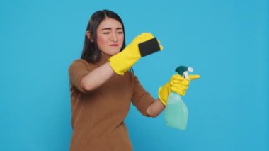 Overwhelmed maid standing in studio over blue background doing intensive cleaning using chemical detergent spray and sponge. Tired housekepper is dedicated to maintaining the cleanliness