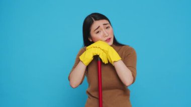 Tired housekepper falling asleep on broom while cleaning client home, being overworked in studio over blue background. Sleepy maid wearing rubber gloves while being was responsible for the houseclean