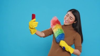 Smiling housekeeper holding colorful feather dust while taking selfie with mobile phone, standing in studio over blue background. Cheerful maid using high-quality detergents and rubber gloves