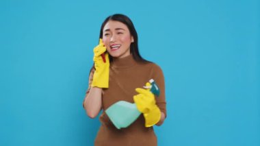 Cheerful housewife wearing protective gloves while holding chemical spray bottle talking at smartphone with remote friend, standing in studio over blue background. Housekeeping and cleaning concept.