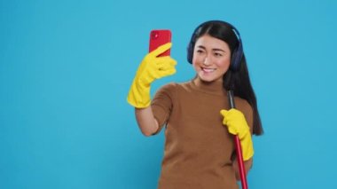 Cheerful housekeeper holding cleaning broom while taking selfie with smartphone, standing in studio over blue background. Smiling maid using high-quality detergents and rubber gloves