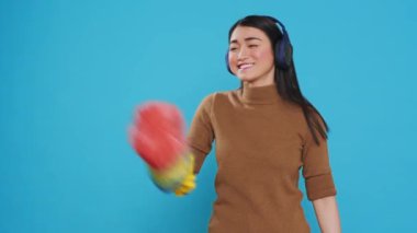 Cheeful housekeeper wearing headphones while dancing and having fun during house cleaning using feather duster. Overwhelmed maid role was vital to the smooth operation and cleanliness of many homes