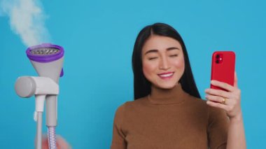 Professional housekeeper ironing clothes using steamer while having videocall meeting on phone, talking with remote friend. Smiling housewife committed to maintaining high standards of cleanliness