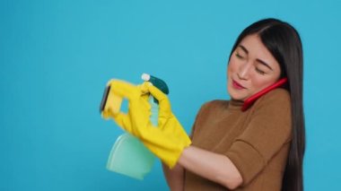 Asian housekeeper using duster and protective gloves to clean house while talking at smartphone with remote friends, having remote discussion. Housekeeping and cleaning concept.