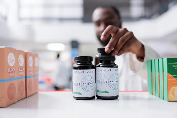 African american pharmacist taking vitamins from drugstore shelf closeup, selective focus on bottles. Pharmacy medic putting pills close view, supplement selling, pharmaceutical service