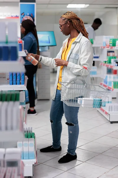 African american buyer choosing sunscreen in drugstore shelf, reading instruction on sunblock cream package. Woman checking skin care lotion in pharmacy store, holding shopping basket