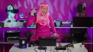 Cheerful performer dancing and interacting with fans in club at nightime, playing techno remix at professional mixer console. Artist with pink hair mixing electronic sounds using audio equipment