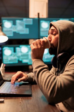 Cyber criminal hacking computer system in office, drinking cup of coffee and acting dangerous. Stealing information from big data software, breaking security server with virus and code.