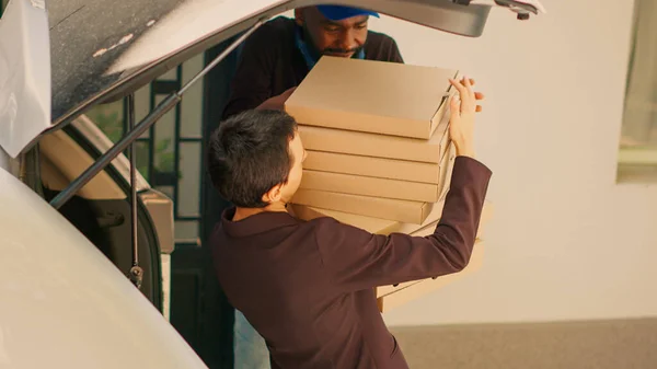 Food delivery worker giving pizza boxes to clients at office entrance, delivering big pizzeria order from car trunk. Taking lunch meal packages out of automobile to deliver takeout. Handheld shot.