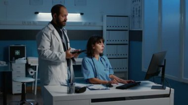 PC 'nin önünde duran asistan, hasta hastalığının teşhisini tartışan sıhhiyeye tıbbi uzmanlığını gösteriyor. Personel gece geç saatlere kadar hastane ofisinde çalışıyor.