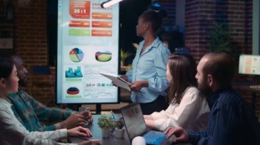 Employee speaking in business meeting presentation, holding report clipboard. Diverse team discussing revenue statistics, planning sales strategy, looking at charts on digital board