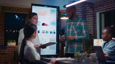 Diverse anaylsts presenting company marketing statistics visualization in business meeting, employees listening to presentation. Office workers in coworking space boardroom at night time