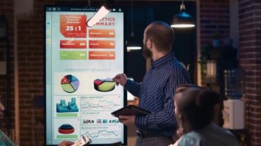 Şirket analisti istatistik şemalarını açıklıyor, iş toplantısında sunum yapıyor, dijital tablet tutuyor. Çalışan, analitik araştırma çizelgelerini gece ekranda gösteriyor. El kamerasıyla..