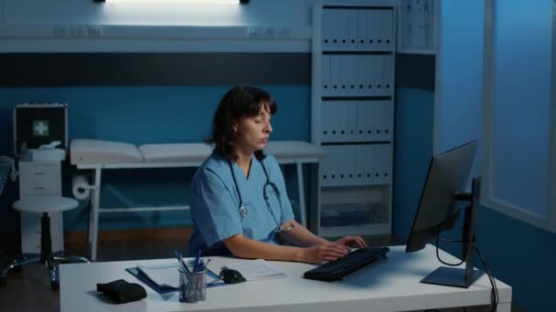 Wyczerpana Pielęgniarka Niebieskim Mundurze Pracująca Godzinami Biurze Szpitalnym Analizująca Doniesienia — Wideo stockowe