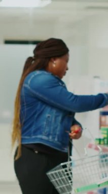 Vertical video: Woman taking medicaments off of drugstore shelves, looking at medical products to buy prescription medicine. Young customer checking leaflets of supplements and pills, retail store.