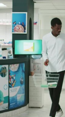 Vertical video: African american customer putting medicaments in basket, looking for vitamins and bottles of pills. Young male client buying prescription treatment and supplements, pharmacy shop.