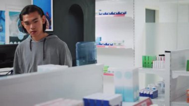 Asyalı genç adam eczane raflarındaki ilaç kutularını analiz ediyor, reçeteli tedavi için vitaminleri inceliyor. Sağlık hizmetleri eczanesinde tıbbi ürünler ve uyuşturucular kontrol ediliyor.