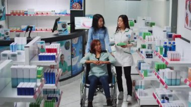 Tekerlekli sandalyedeki kadın hademe ve kadın, ilaç ya da haplar hakkında uzman soruyorlar, eczanede reçeteli ilaç arıyorlar. Eczanede engelli kişilere yardım eden sosyal hizmet görevlisi.