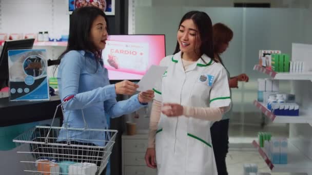 薬局の従業員は アジアのクライアントに薬や薬を与えるために処方紙を読んで ドラッグストアの女性に助言を与えます 薬や薬の箱の話をしている人たちは — ストック動画
