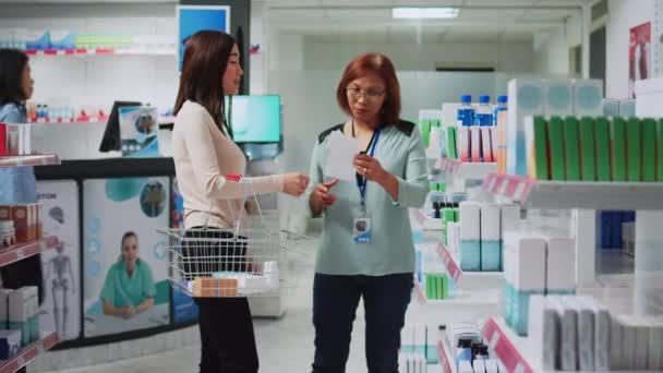 医薬品やビタミンを顧客に与えるために薬局で処方紙を読んで若い薬剤師 医薬品店からサプリメントや薬箱を購入するアジア人 — ストック動画