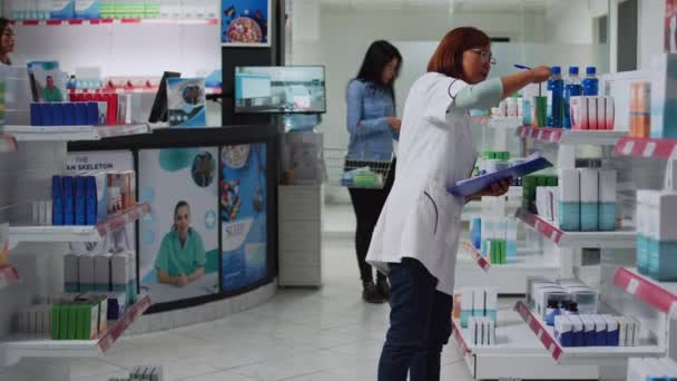 アジアの薬剤師は 医薬品在庫に関するメモを取り 顧客に販売するために紙で薬在庫に取り組んでいます ドラッグストアの従業員は ビタミンの薬やボトルの箱を見て — ストック動画