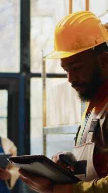 Dikey video: Depo çalışanı, depoda envanter sayımı yapmak için tarayıcı ve dijital tablet kullanarak, barkodları depoda tarıyor. Depoda raflarda ürünlerle çalışan genç bir çalışan..