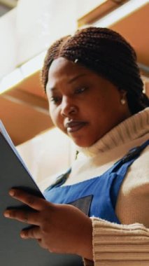 Dikey video: Depoda stok lojistiği üzerinde çalışan kadın çalışan envanter listesini kontrol ediyor. Ürünlerin dağıtımını hazırlamak için perakende satış yapan genç bir çalışan. El bilgisayarı