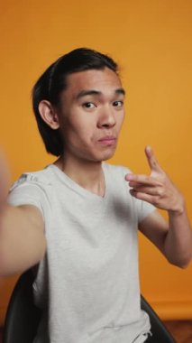 Dikey video: Asyalı gülümseyen adamın kamera önünde fotoğraf çekerken görüntüsü, turuncu arka planda komik ve aptalca davranıyor. Stüdyoda fotoğraflara poz veren, gülümseyen ve mutlu olan neşeli gündelik modeller. Kaygısız