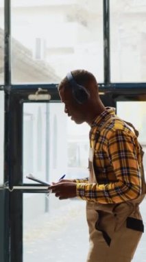 Dikey video: Depoda dans eden genç yetişkin tuhaf davranıyor, kulaklıkla neşeli şarkılar söylüyor. Modern depo çalışanı dans figürleri sergiliyor ve rafların yanında kutularla aptalca davranıyor..