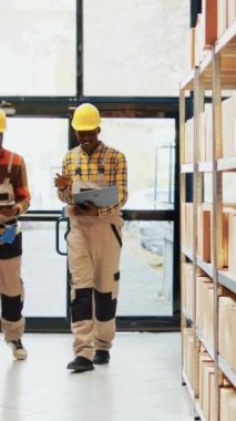 Dikey video: Stok envanterini planlamak için dizüstü bilgisayar ve lojistik listeleri kullanan erkek işçiler, palet ve kutulara bakıyorlar. Depo çalışanları dağıtım için malzeme hazırlıyor.